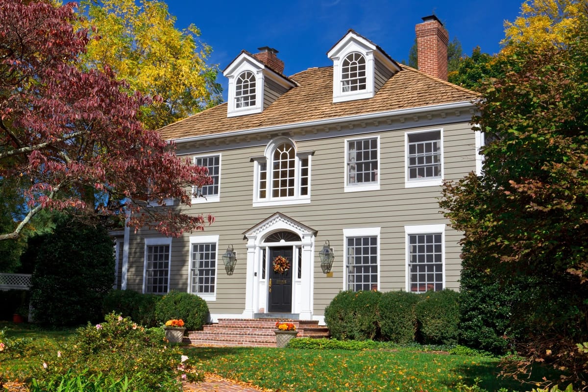 A two-story house with gray siding, one of various house siding options, white trim, and a wooden shingle roof. It features two chimneys and several windows with shutters. The yard is landscaped with bushes and autumn trees, and a wreath decorates the front door.