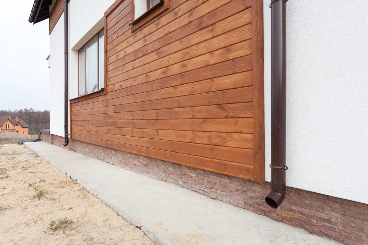 Side view of a house with light plaster and horizontal wooden siding, reminiscent of guides on how to clean wood siding. A brown metal downspout runs to the concrete pathway below. In the grassy background, a few houses are visible under the clear sky.