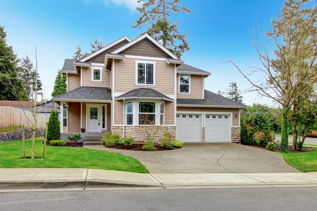 A two-story house with a light brown exterior and gray roof boasts well-maintained siding. It features a front porch, a driveway leading to a double garage, and is surrounded by lush trees and shrubs. The sky above is clear and blue.