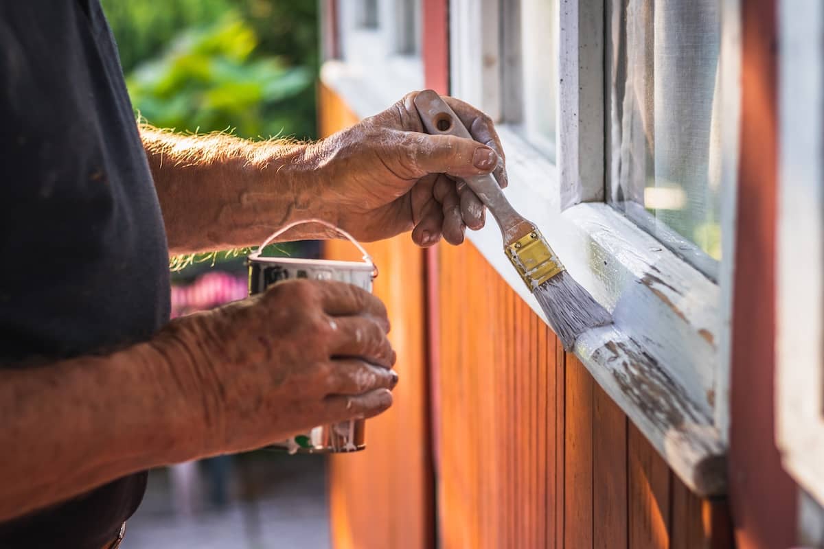 A person is painting the exterior frame of a window white with a paintbrush. The surface is wooden.