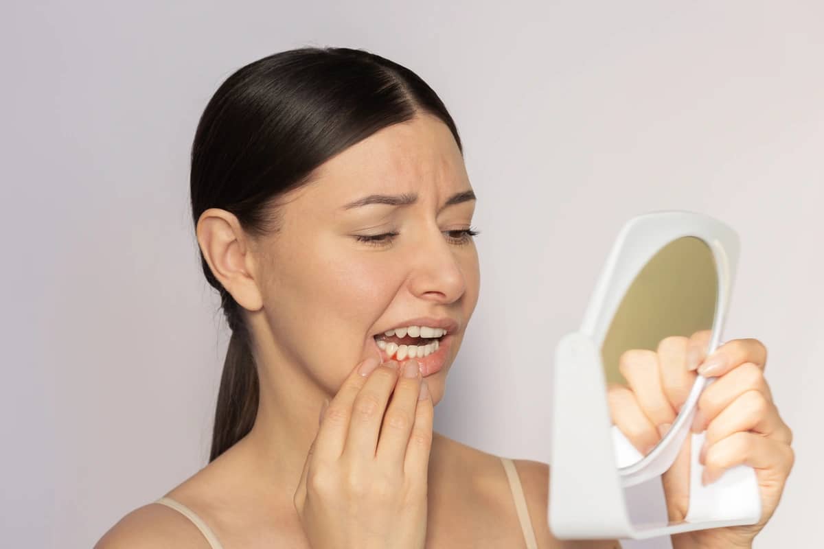 A woman with long dark hair looks concerned as she examines her teeth in a small round mirror. Her other hand touches her cheek, suggesting discomfort. The background is a plain light color.