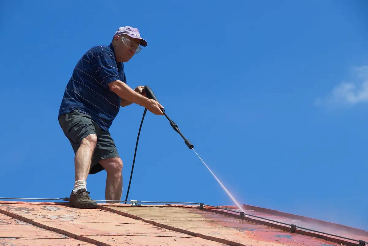 An older man in a striped shirt, shorts, and a cap uses a pressure washer to clean a sloped roof under a clear blue sky.