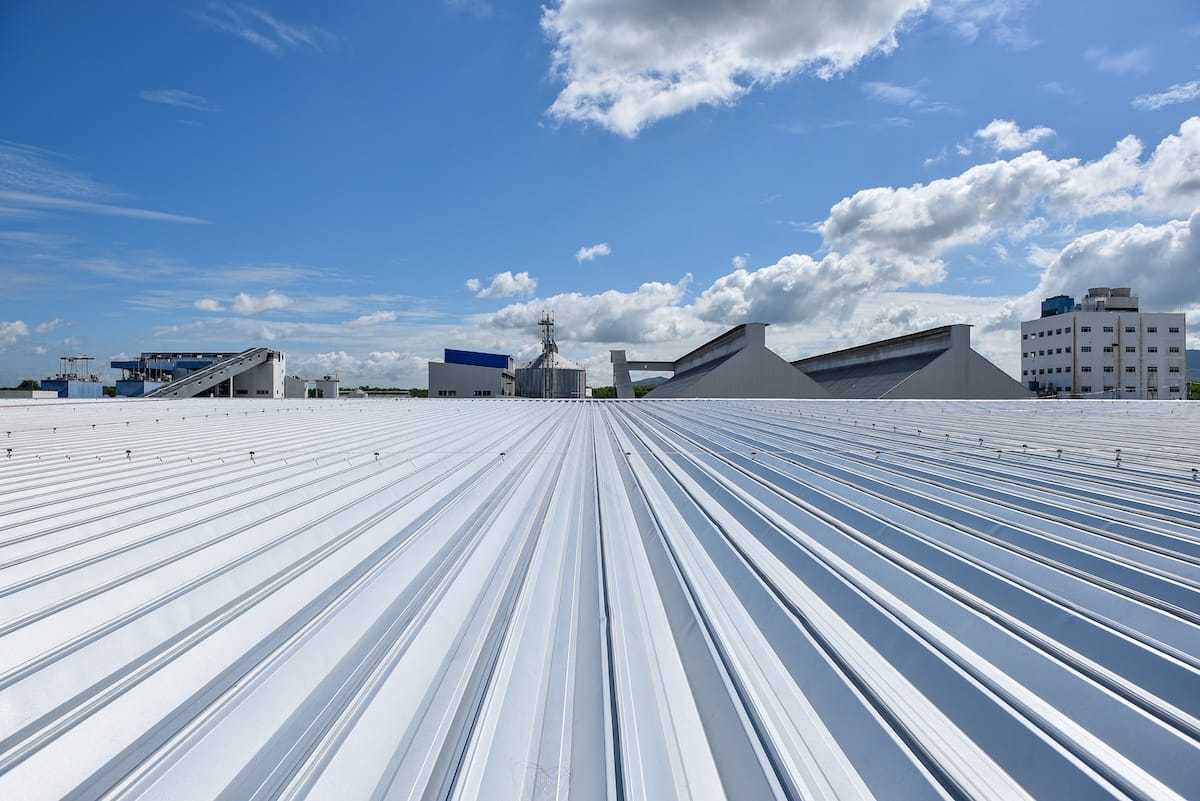 Wide-angle view of a shiny, flat metal roof under a blue sky with scattered clouds, featuring various factory buildings and equipment in the background.