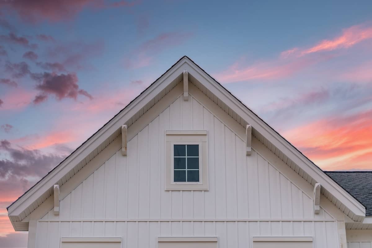A white house gable with vertical siding is set against a vibrant sky. The sky features a blend of pink, blue, and purple hues during sunset. A small, central window sits beneath the peaked roof, which has simple detailing.