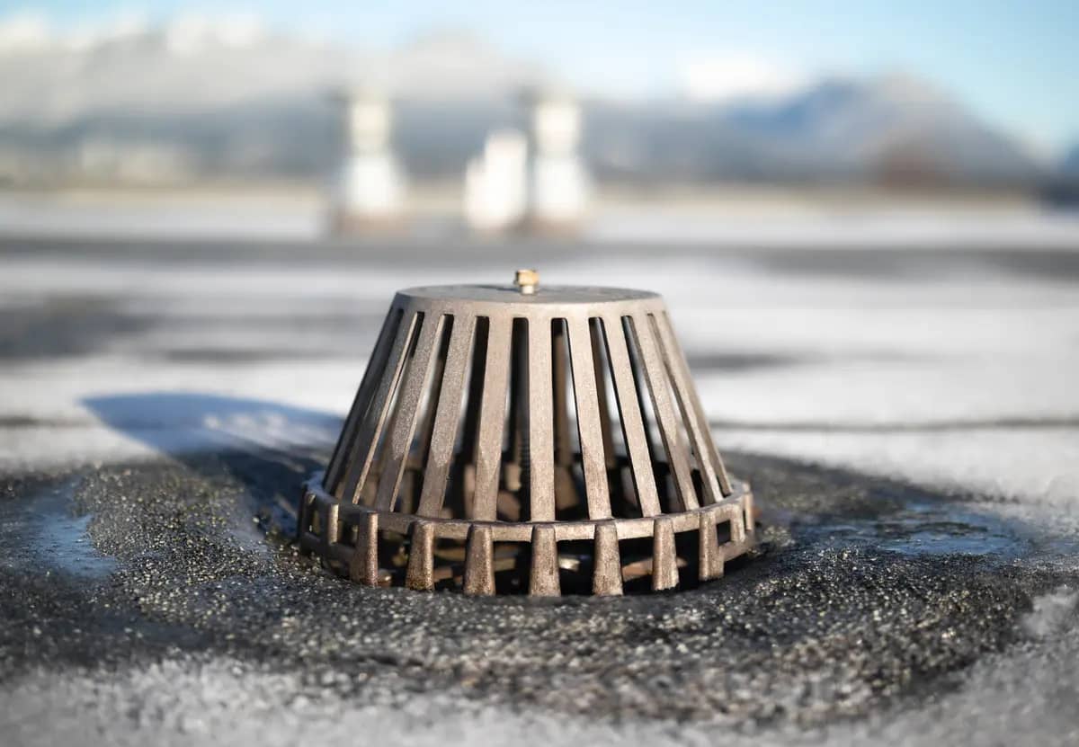 Close-up of a metal roof drain surrounded by melting snow on a flat surface. The drain's conical structure is designed to prevent debris from clogging. The background shows blurred mountains under a clear blue sky.