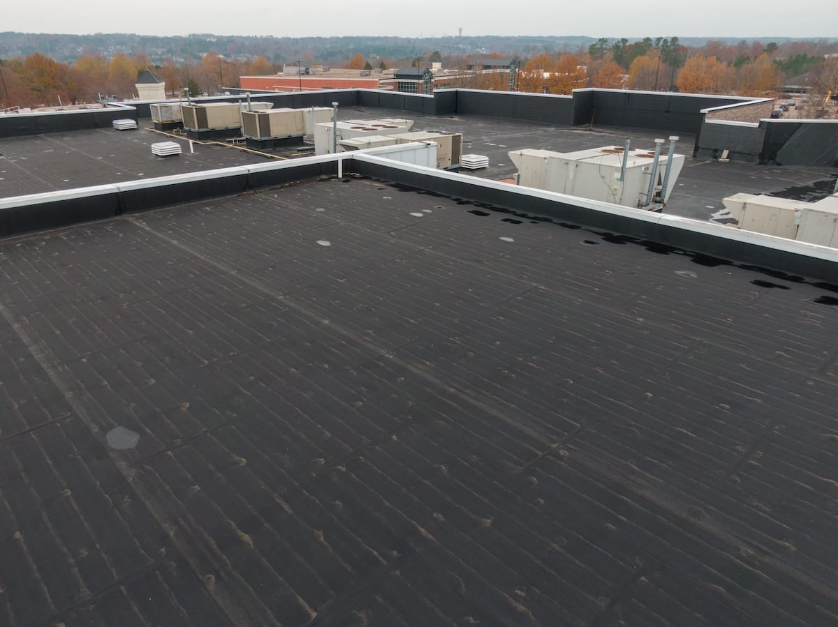 A flat rooftop with black surface materials and several HVAC units and vent pipes. Trees with autumn foliage are visible in the background under a cloudy sky.