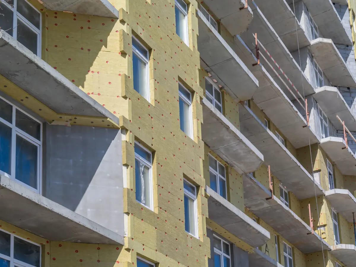 Close-up of a building under construction with exposed yellow insulation. Several floors with balconies are visible. The building features large windows and concrete floors. The sun casts shadows, highlighting the texture of the materials.