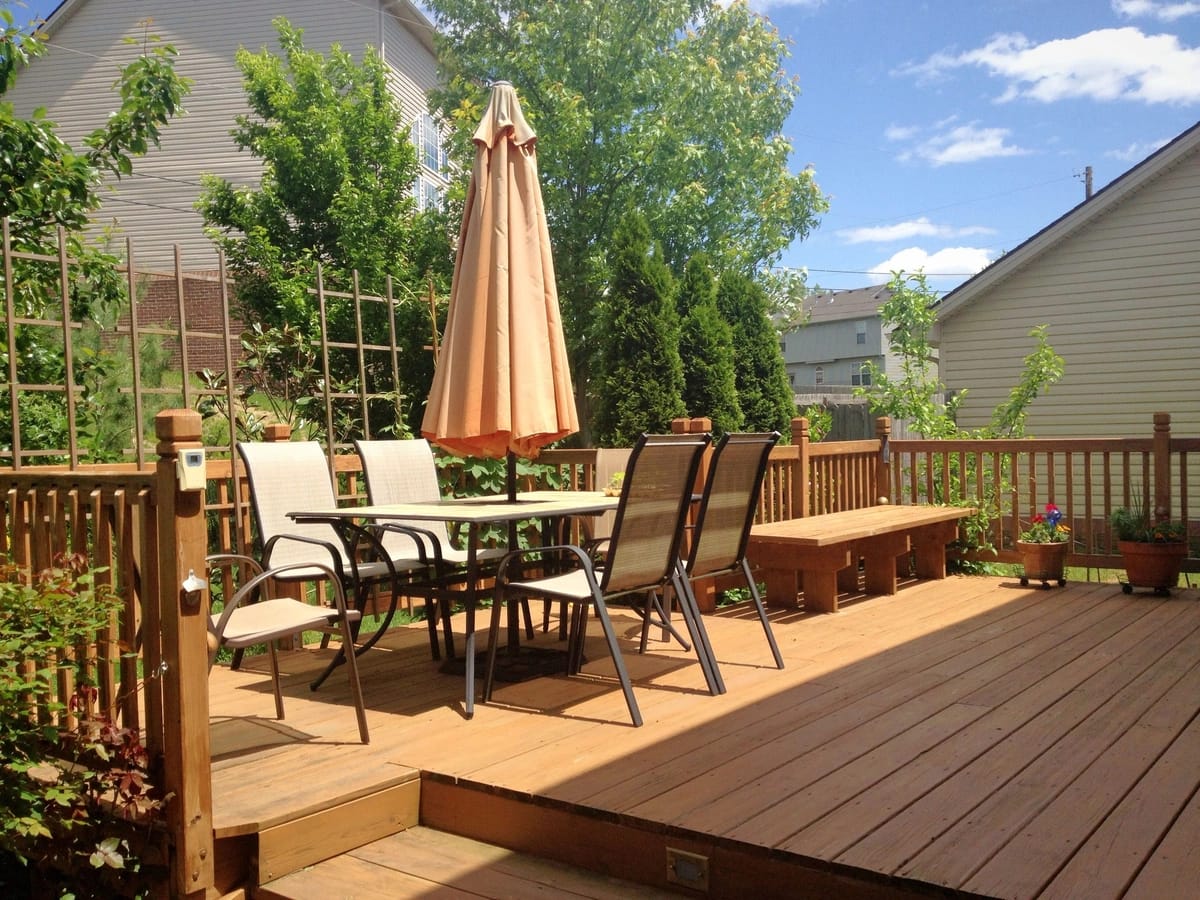 A sunny backyard deck with a wooden table and six chairs, shaded by a beige umbrella. Potted plants and a wooden bench enhance the space, while green foliage frames neighboring houses under a blue sky. Recently finished with expert deck repair, it invites you to relax in style.
