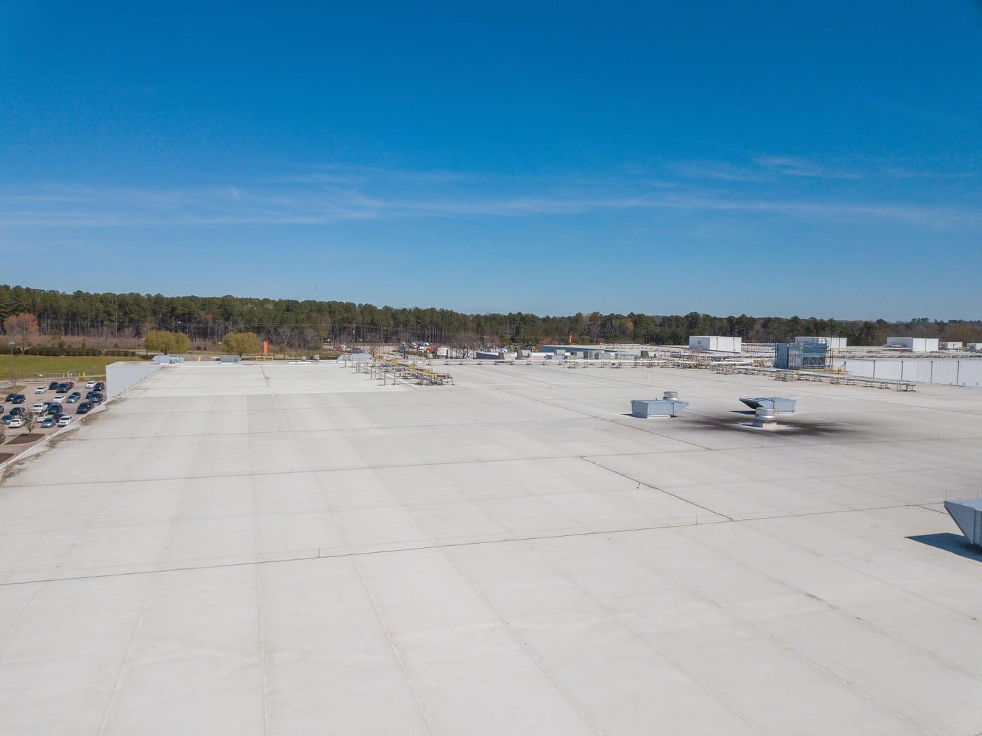 Aerial view of a large, flat industrial rooftop under a clear blue sky. The rooftop features air conditioning units and skylights. In the background, there are trees and some industrial buildings.