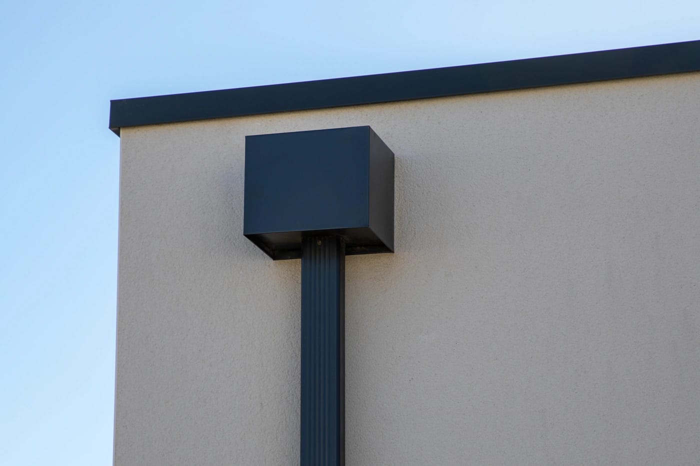 A minimalist architectural detail featuring a square, black metal box affixed to the upper corner of a beige building. A vertical black pipe extends downward from the box against a clear blue sky.