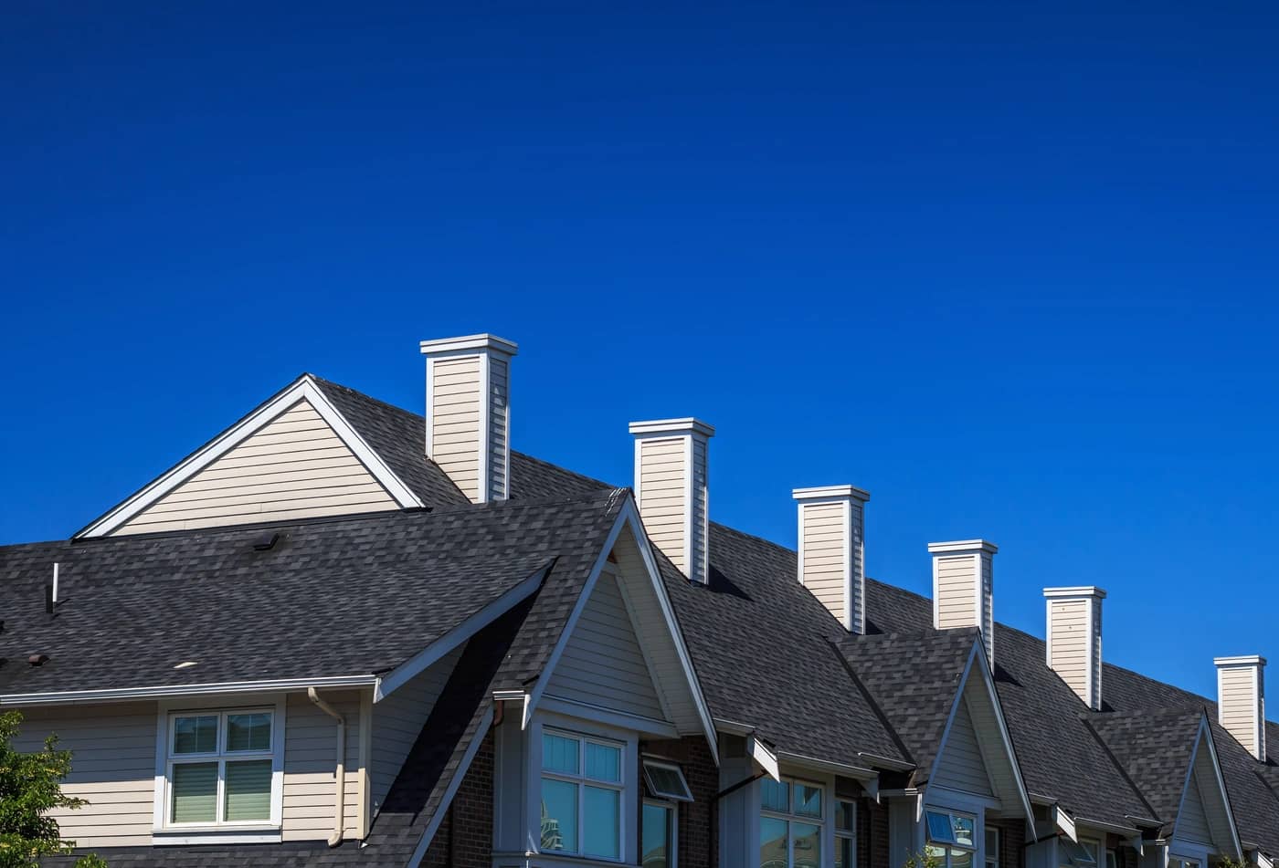 A row of townhouses with dark gray roofs and multiple chimneys against a clear blue sky. The houses feature light-colored siding and triangular gables.