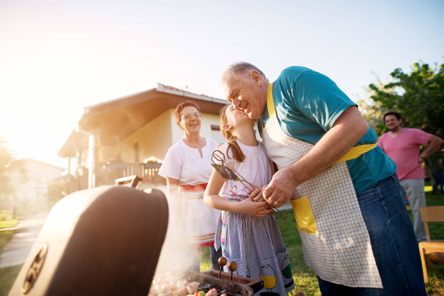 A joyful family scene unfolds in a backyard with an elderly man wearing an apron, affectionately hugging a young girl while holding a skewer. Beside them, an older woman smiles warmly. In the background, near the house with commercial roofing, a man tends to food grilling in the foreground.