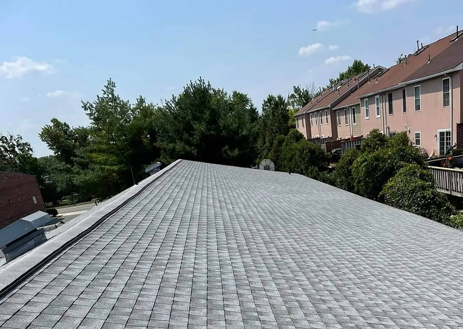 View of a residential rooftop covered with gray shingles, stretching towards a line of townhouses on the right. Trees and a clear blue sky are in the background.