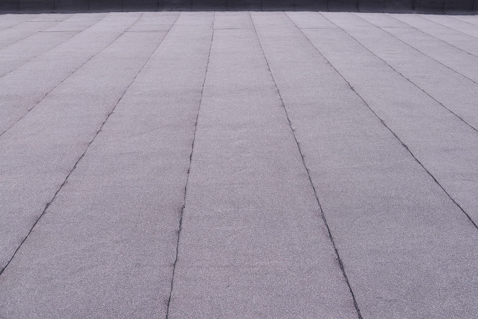 A wide view of a flat rooftop with a uniform, gray gravel-like surface. The roof is segmented by lines running parallel, creating a series of long rectangular sections. The background shows a darker border at the roof's edge.