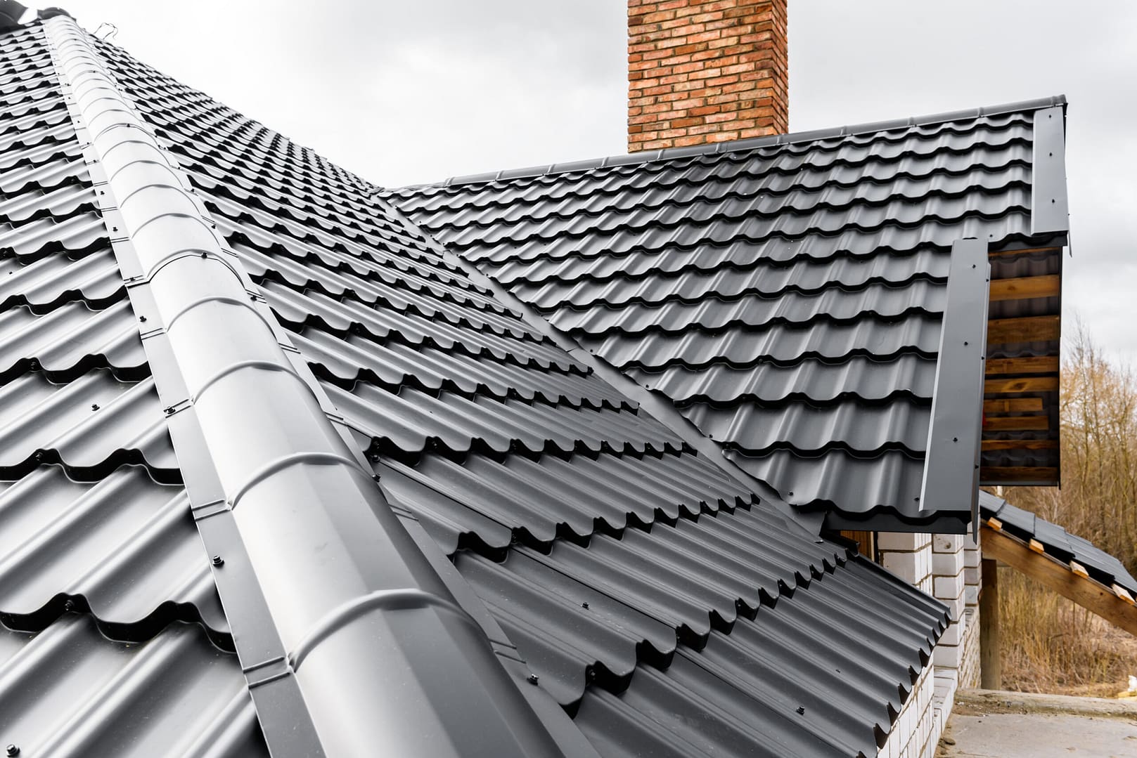 Close-up of a house's roof with sleek black metal roofing tiles. The roof features a brick chimney, and part of the overhang is visible.