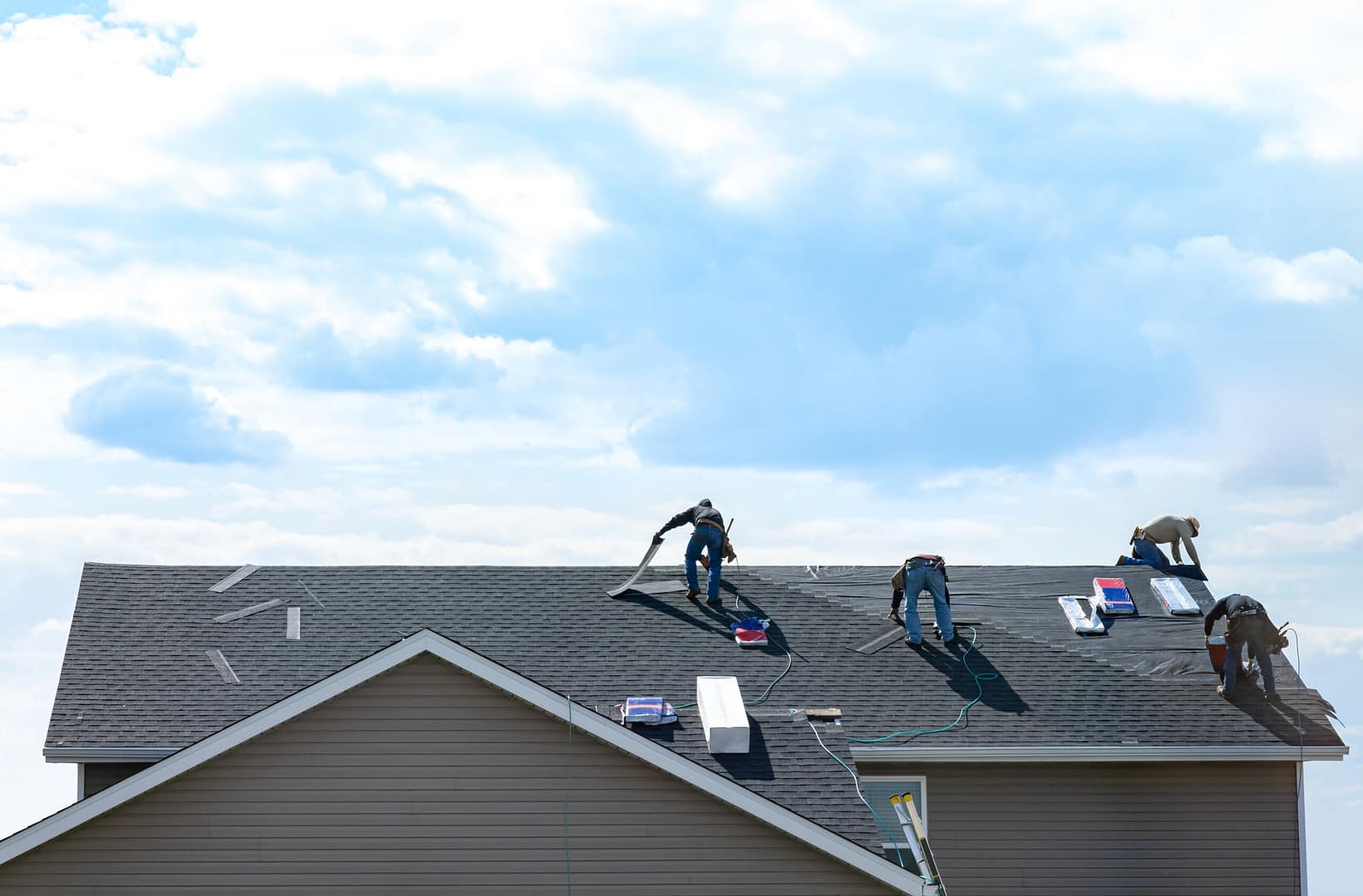 Four workers are installing shingles on a house roof under a bright, cloudy sky as roofing materials and tools are spread out, one wonders how long does a 30 year roof really last
