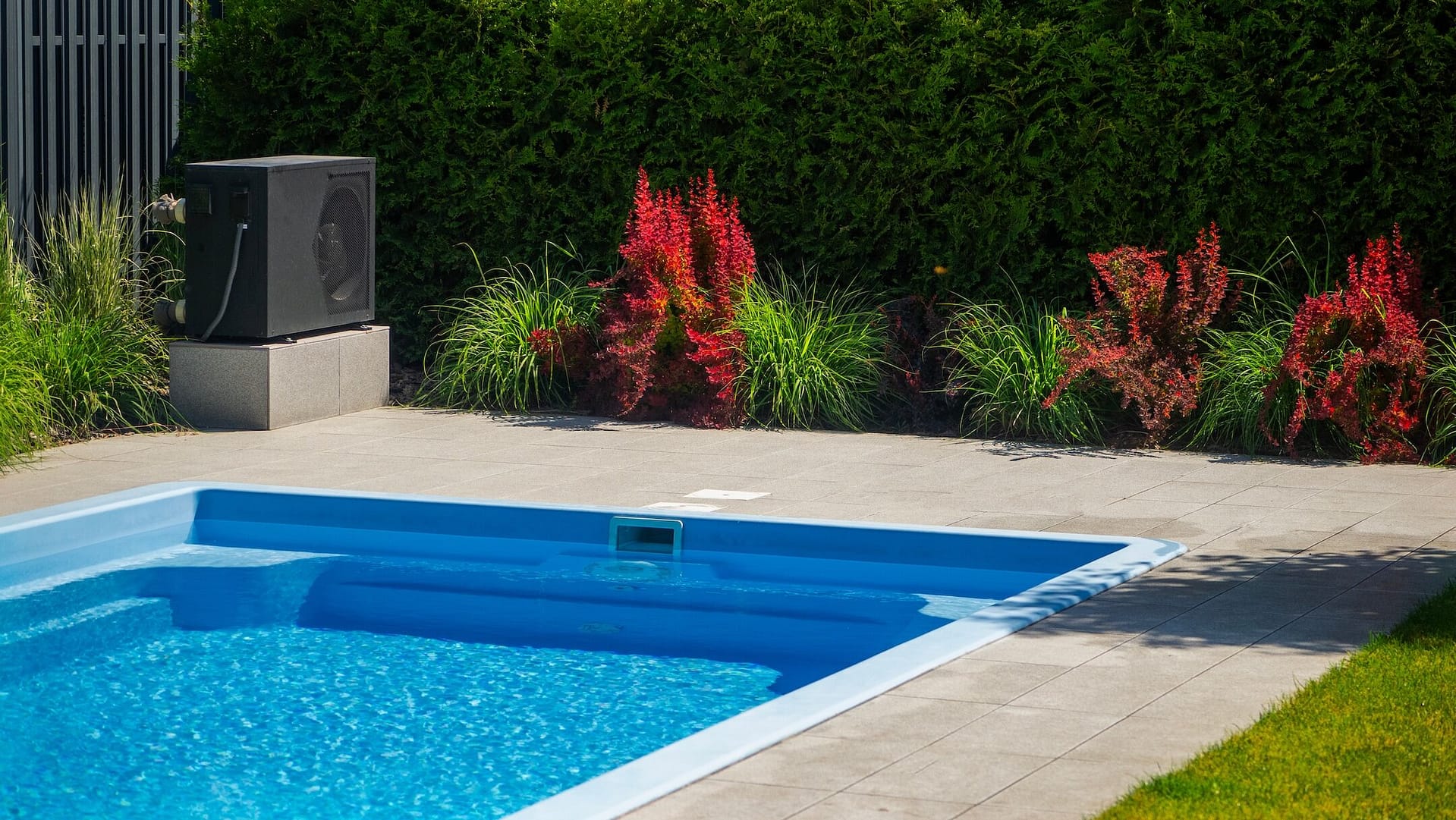 An outdoor heat pump unit is situated next to a swimming pool, mounted on a concrete platform. The surrounding area features decorative plants, including tall grasses and neatly maintained bushes, enhancing the visual appeal. The environment is clean and well-kept, with the blue pool water offering a refreshing contrast to the greenery. The modern heat pump and the beautifully landscaped garden create a harmonious and functional outdoor setting.