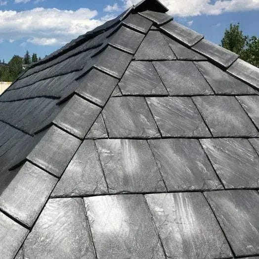 Close-up view of a roof with overlapping gray slate tiles and rubber shingles beneath a clear blue sky. The tiles have a rough texture, showcasing their natural stone appearance. Trees and part of a building are visible in the background.