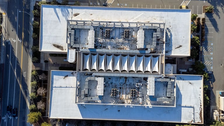 Aerial view of a large, rectangular industrial building with a white roof. The roof features two sections of ventilation or HVAC systems and a central row of triangular, tent-like structures. Surroundings include parked cars and trees.