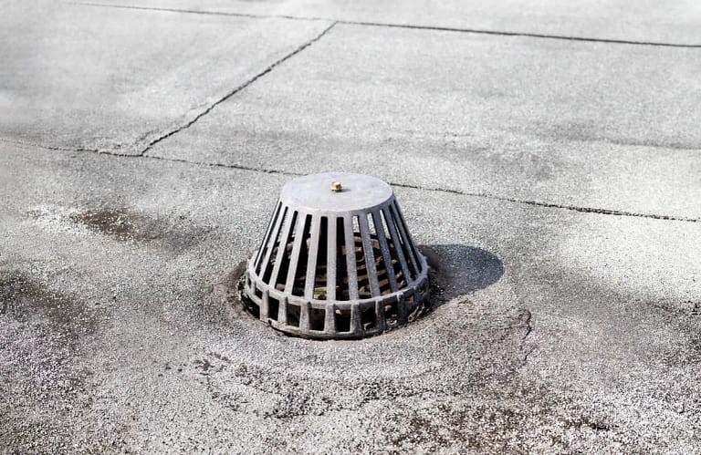 A metal storm drain cover is situated on a flat, textured concrete surface. The drain cover is circular with vertical slats and casts a shadow on the ground. Subtle cracks and variations in the surface texture are visible.