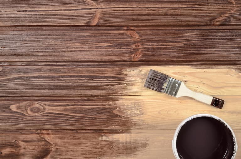 A paintbrush and can of deck stain rest on wooden planks, with half of the surface stained and the other half still light