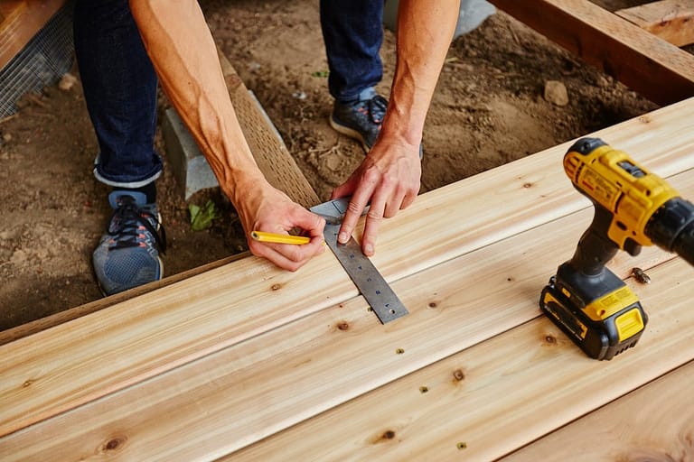 A person measures and marks a wooden plank during deck installation with a ruler and pencil