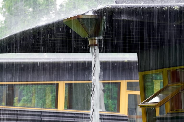 Close-up of a roof with rainwater cascading down a chain from a triangular downspout. The building has large windows with yellow frames. The surrounding trees are blurred by the rain.