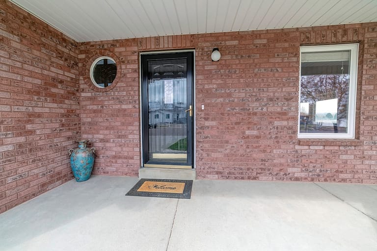 House entrance with glass storm door and front door against red brick wall. Round and rectangular window window can be seen by the porch with decorative jar.