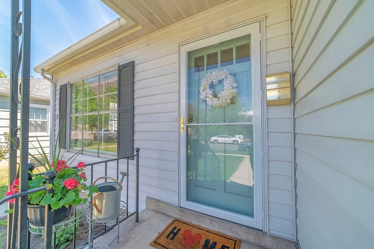 Front view of house on real estate listing with glass storm door and green door. Porch with railing, window with shutters, and white wall siding can be seen at the home exterior.