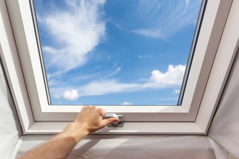 A hand reaching to open a skylight with a view of a blue sky and clouds