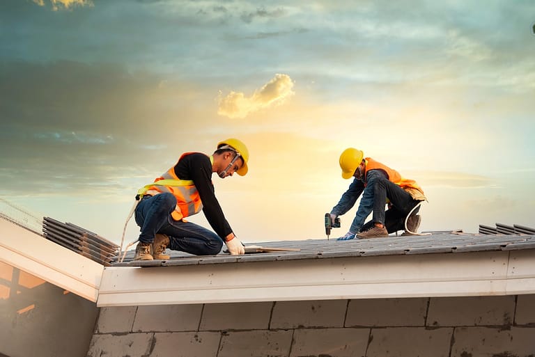 Two construction workers in safety vests and helmets install roofing materials on a house at sunset