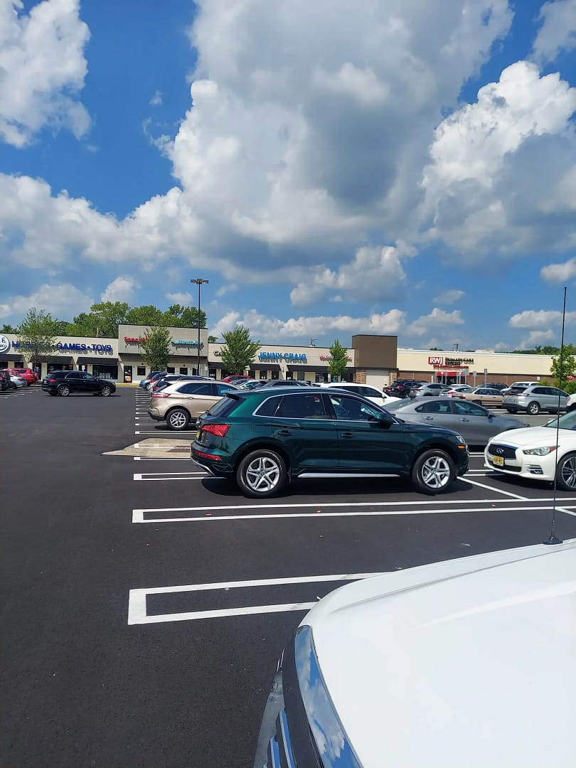 A parking lot with several parked cars under a partly cloudy sky. In the background, there are retail stores, including GameStop and AT&T. The surrounding trees add greenery to the scene.