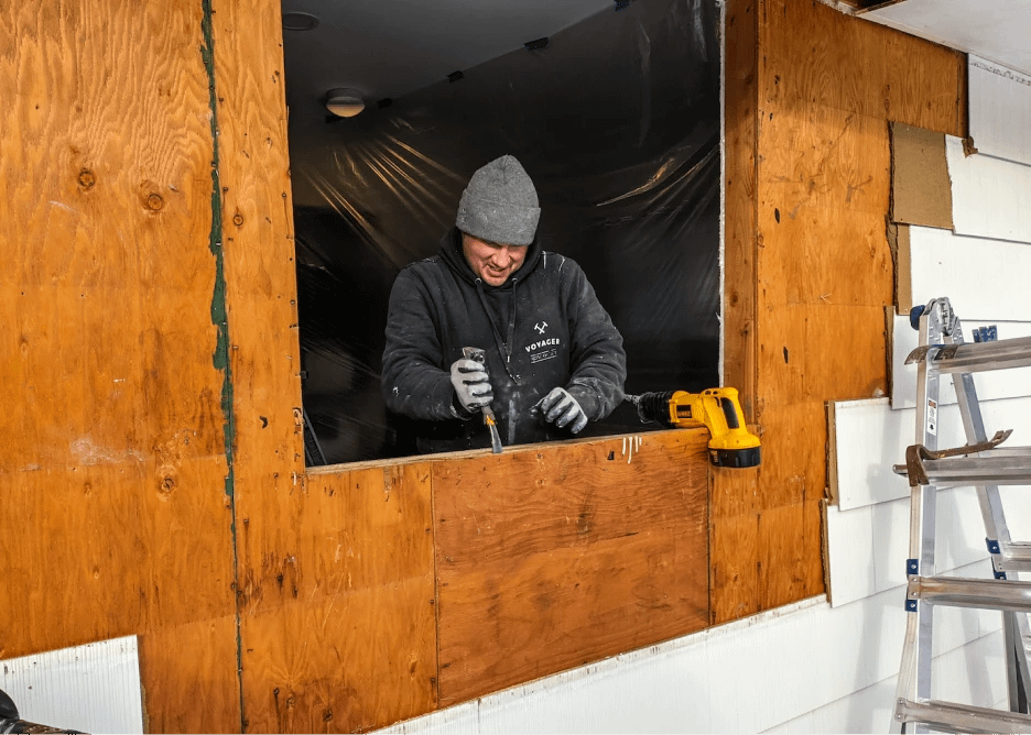 A person in a gray beanie and black hoodie works on a large wooden board attached to a wall, using a power tool. A yellow drill lies nearby. There's a stepladder on the right. Plastic sheeting covers the background.