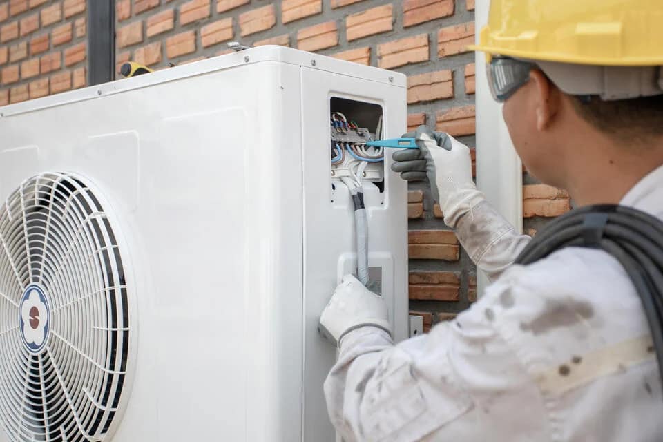 heat-pump-repair-tech-wearing-safety-hat-performing-maintenance-on-heat-pump-near-brick-sided-house