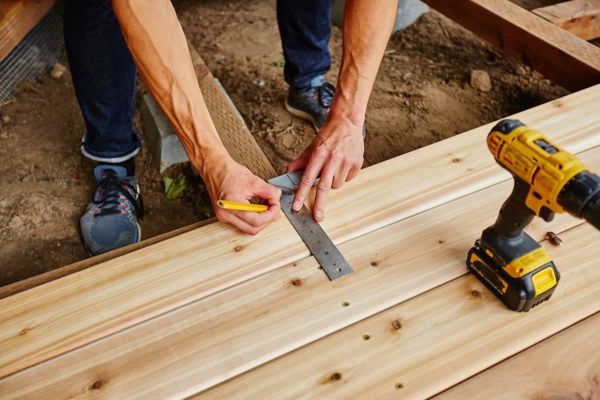 A person measures and marks a wooden plank during deck installation with a ruler and pencil