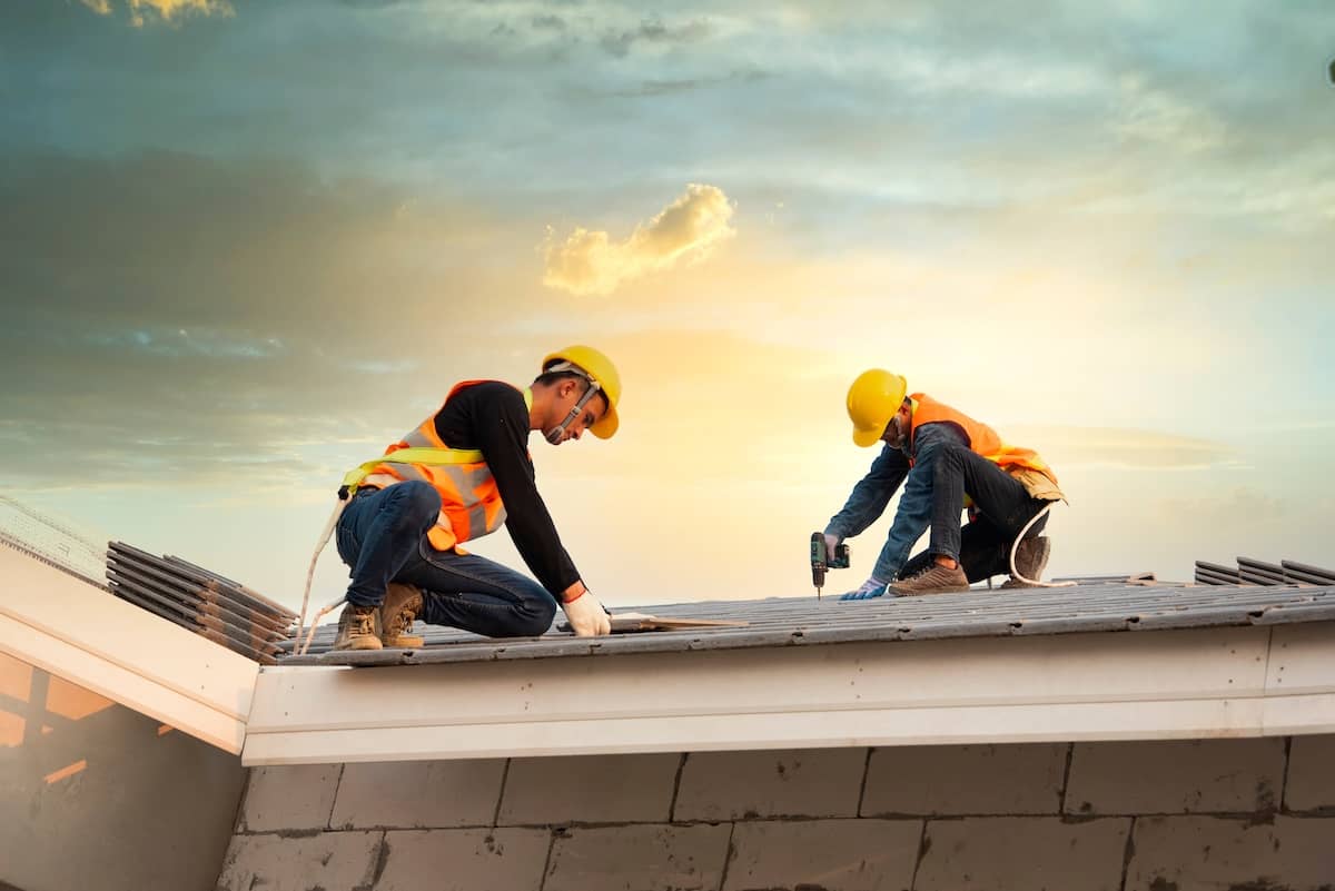 Two construction workers in safety vests and helmets install roofing materials on a house at sunset