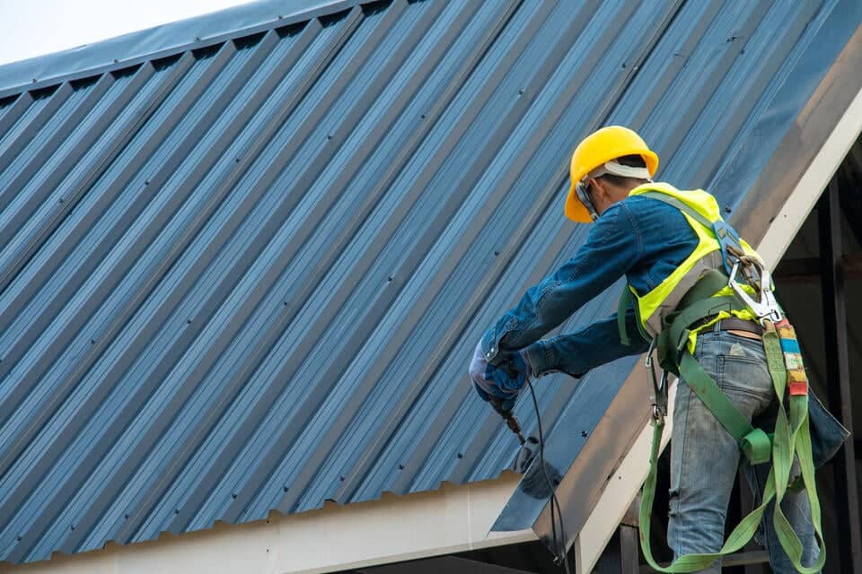 metal roof cost-roofer in hard hat and safety vest and harness installing metal roof panels on a house