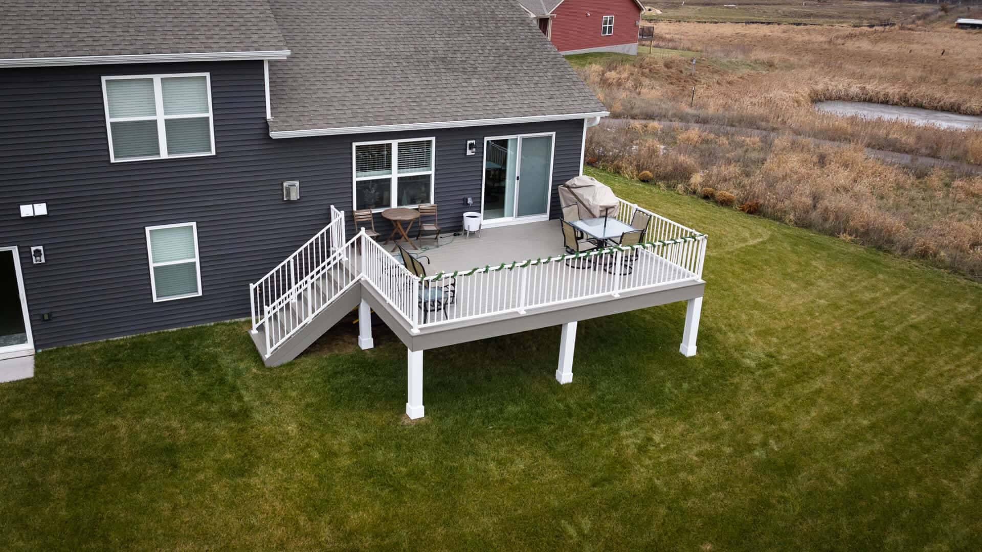 Aerial view of a house with a dark gray exterior and white-framed windows. The house features a white-railed, elevated deck with stairs leading down to a green lawn. The countryside landscape surrounds the property.